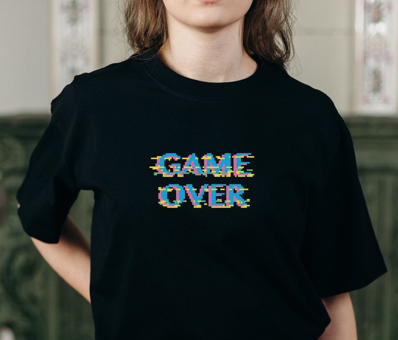 Op de afbeelding: Zwart T-shirt met een gepixelde "GAME OVER" afbeelding in blauw, roze en geel.