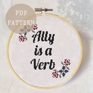 以下が含まれることがあります： 「Ally is a Verb」と刺繍された白い刺繍枠。黒い糸で刺繍されており、小さな赤、青、緑の花で囲まれています。