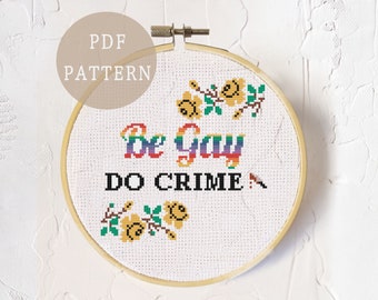 May include: A cross-stitch pattern featuring the text "Be Gay Do Crime" in rainbow colours with yellow flowers on either side. The pattern is displayed in a wooden embroidery hoop.