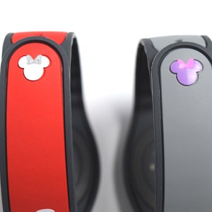 May include: Two MagicBand wristbands. One is red with a white Minnie Mouse head and silver bow. The other is gray with a purple Minnie Mouse head and bow. Both have a black border and are on a white background.