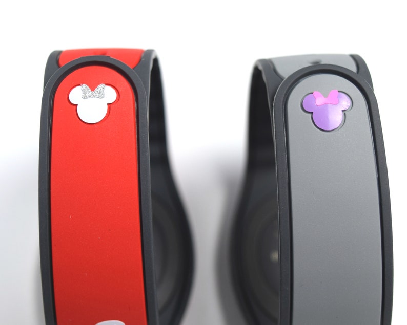 May include: Two MagicBand wristbands. One is red with a white Minnie Mouse head and silver bow. The other is gray with a purple Minnie Mouse head and bow. Both have a black border and are on a white background.