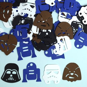 May include: A collection of die-cut felt shapes featuring iconic science fiction character silhouettes. The assortment includes brown Wookiee heads, black Darth Vader masks, white Stormtrooper helmets, and blue R2-D2 droid shapes scattered across a light blue surface.