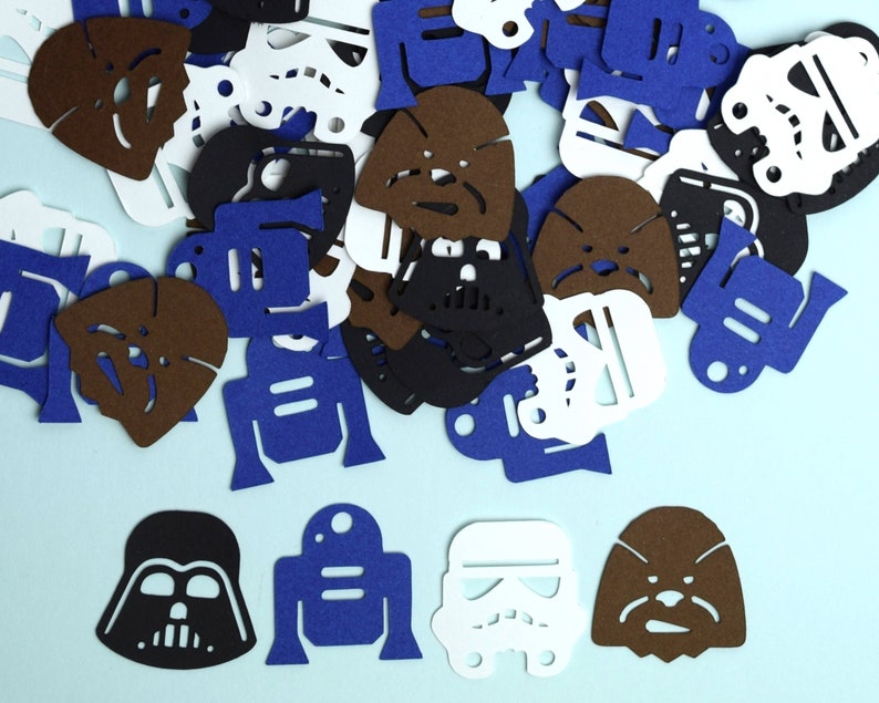May include: A collection of die-cut felt shapes featuring iconic science fiction character silhouettes. The assortment includes brown Wookiee heads, black Darth Vader masks, white Stormtrooper helmets, and blue R2-D2 droid shapes scattered across a light blue surface.