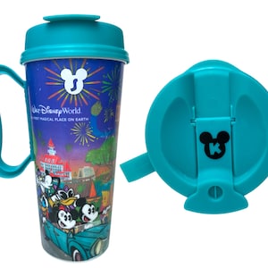 May include: A teal travel mug with a handle and a matching lid featuring a black Mickey Mouse icon. The mug displays a colourful Walt Disney World graphic with fireworks, a car, and characters. The lid has a sliding closure with a black Mickey icon.