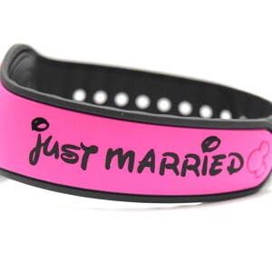 May include: A bright pink silicone wristband with a black interior and a series of adjustment holes. The text "JUST MARRIED" is printed in black on the outer surface, accompanied by a small black mouse ear icon at the end of the text.
