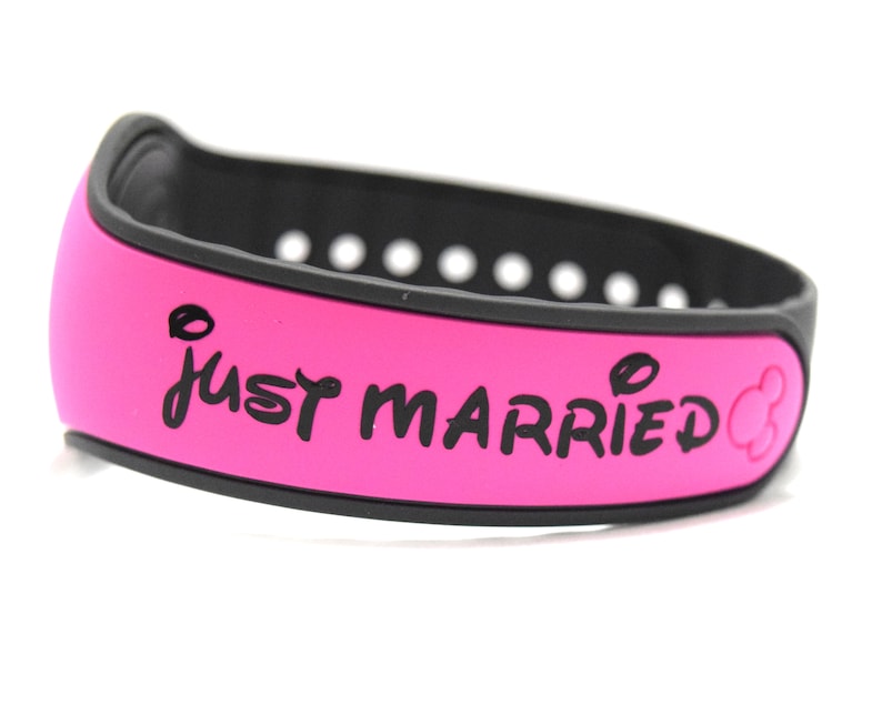 May include: A bright pink silicone wristband with a black interior and a series of adjustment holes. The text "JUST MARRIED" is printed in black on the outer surface, accompanied by a small black mouse ear icon at the end of the text.