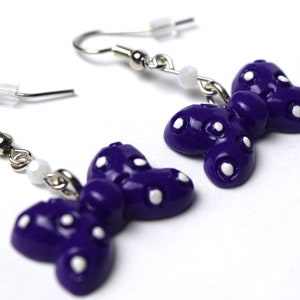 以下が含まれることがあります： 白い水玉模様の紫色の蝶のイヤリング。イヤリングはポリマークレイ製で、銀色のフックが付いています。