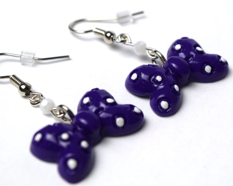 以下が含まれることがあります： 白い水玉模様の紫色の蝶のイヤリング。イヤリングはポリマークレイ製で、銀色のフックが付いています。