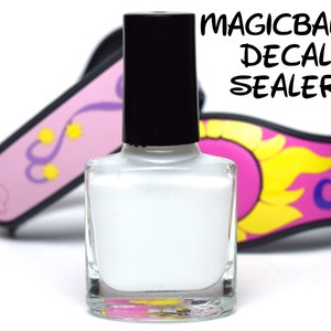 Może przedstawiać: Butelka białego Magicband Decal Sealer. Butelka jest przezroczysta i ma czarną zakrętkę. Butelka stoi na białym tle.