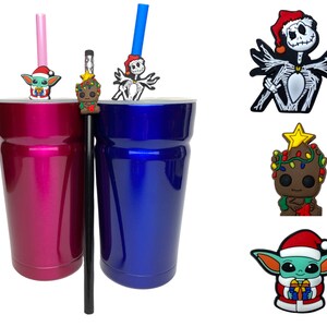 May include: Two stainless steel tumblers, one pink and one blue, with various Christmas-themed character straw toppers. The characters include a green alien, a green tree, a skeleton, and a brown alien.