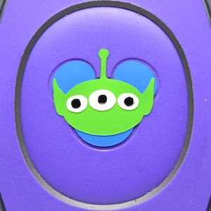 May include: A purple and black MagicBand featuring a green alien character with three eyes and blue Mickey Mouse ears. The alien is centered on a purple circular design.