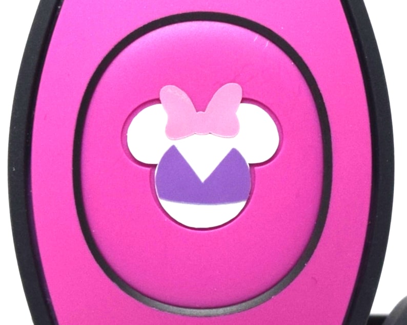 Op de afbeelding: Een roze plastic bedel met een zwarte omtrek. De bedel heeft een wit silhouet van een cartoonmuis die een paarse badpak en een roze strik draagt.