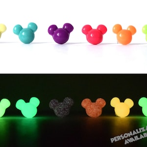 May include: Eight glow-in-the-dark Mickey Mouse head shaped buttons in various colours, including yellow, green, purple, pink, orange, and red. The buttons are shown in two rows, with the top row showing the buttons in their regular colours and the bottom row showing the buttons glowing in the dark.