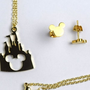 Puede incluir: Conjunto de collar y pendientes de color dorado con la silueta de un castillo con las orejas de Mickey Mouse recortadas. El collar tiene una cadena delicada y los pendientes son de estilo de bot&oacute;n.