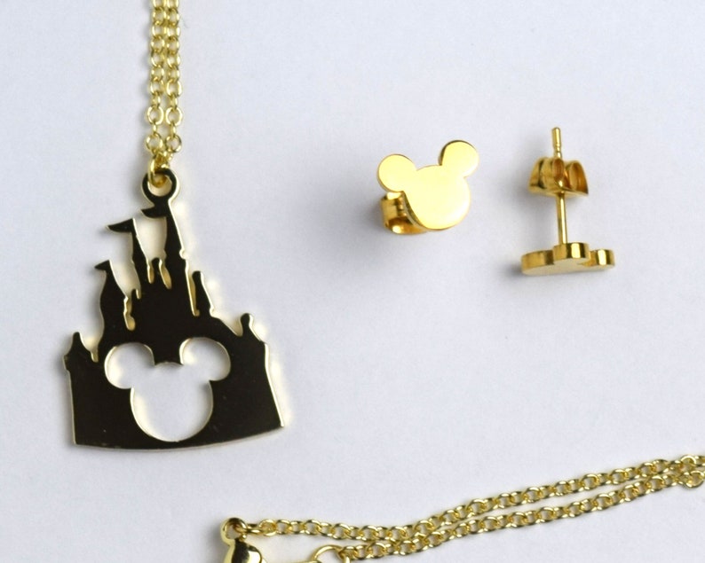 Puede incluir: Conjunto de collar y pendientes de color dorado con la silueta de un castillo con las orejas de Mickey Mouse recortadas. El collar tiene una cadena delicada y los pendientes son de estilo de bot&oacute;n.