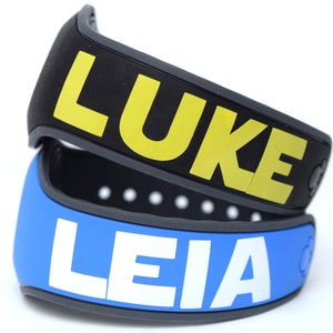 Pode incluir: Duas pulseiras de silicone pretas e cinzas com os nomes "LUKE" em amarelo e "LEIA" em branco.