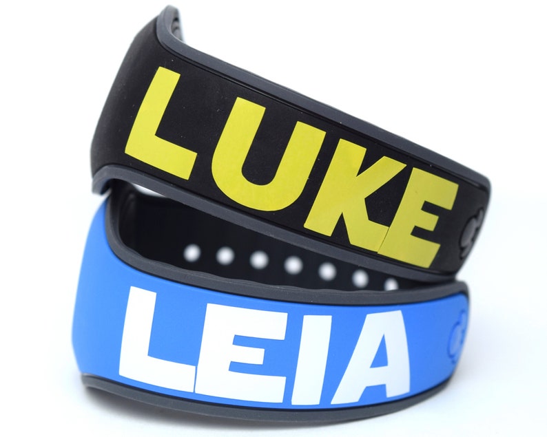Pode incluir: Duas pulseiras de silicone pretas e cinzas com os nomes "LUKE" em amarelo e "LEIA" em branco.