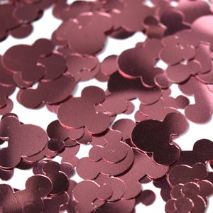 May include: A close-up of rose gold confetti shaped like Mickey Mouse heads. The confetti is scattered on a white background.
