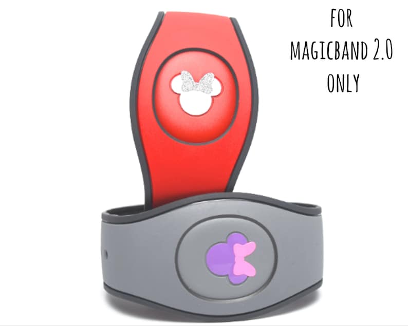 May include: Two MagicBand 2.0 accessories. One is red with a silver glitter Minnie Mouse design, the other is gray with a purple bow design. The text "FOR MAGICBAND 2.0 ONLY" is visible.