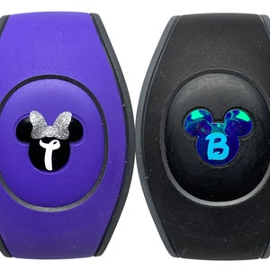 May include: Two purple and black Disney MagicBands with Mickey Mouse ears. The purple band has a silver glitter bow on Mickey's head and a white letter T inside the ears. The black band has a blue and green iridescent Mickey Mouse head with a blue letter B inside the ears.