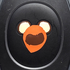 May include: A black circular button with a cartoon orange bear face with brown eyes and a brown mouth. The bear face has two white circles for ears.