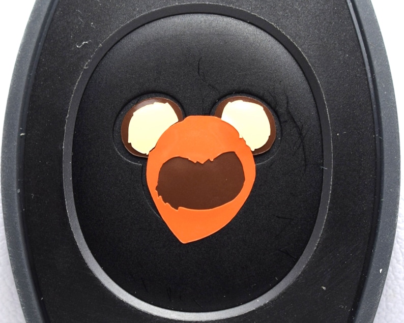 May include: A black circular button with a cartoon orange bear face with brown eyes and a brown mouth. The bear face has two white circles for ears.