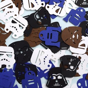 May include: A collection of die-cut paper shapes featuring iconic science fiction character silhouettes. The assortment includes white stormtrooper helmets, black Darth Vader masks, brown Wookiee faces, and blue droid figures scattered across the frame.