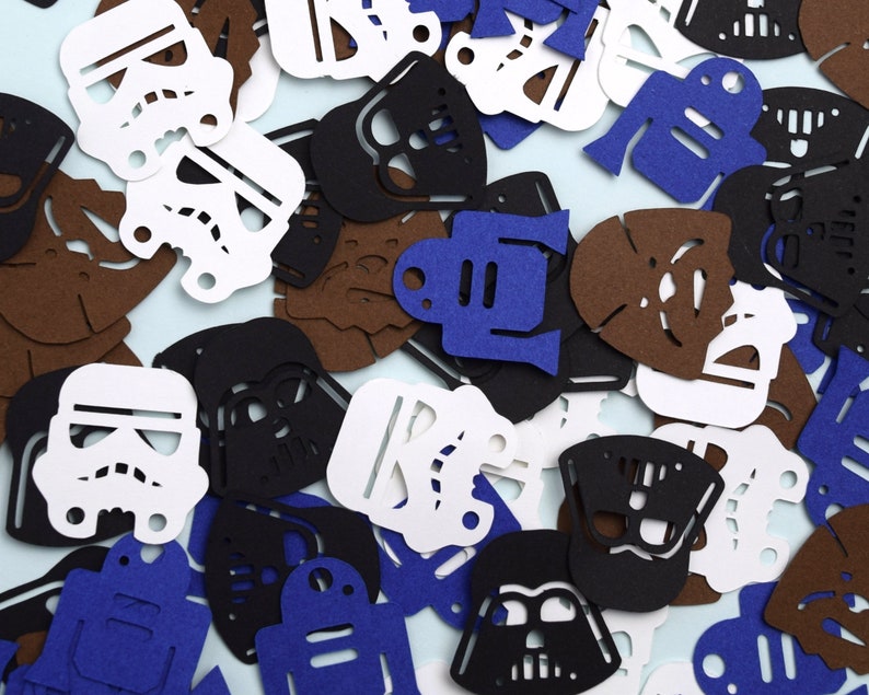 May include: A collection of die-cut paper shapes featuring iconic science fiction character silhouettes. The assortment includes white stormtrooper helmets, black Darth Vader masks, brown Wookiee faces, and blue droid figures scattered across the frame.