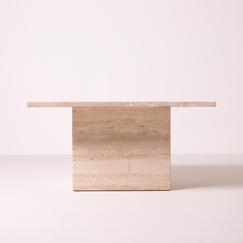 Pu&ograve; includere: Un tavolino rettangolare in pietra beige con un piano orizzontale che si estende da una base solida. Il tavolo ha un design minimalista e un aspetto naturale e strutturato, adatto agli spazi abitativi moderni.