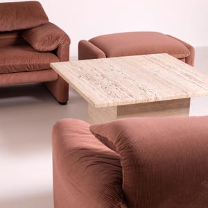 Pu&ograve; includere: Un salotto con un tavolino quadrato in travertino e due poltrone in velluto a coste color rosa tenue. &Egrave; presente anche un pouf nello stesso tessuto e colore. L'arredamento &egrave; disposto su uno sfondo bianco.