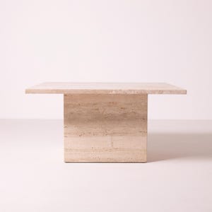 Pu&ograve; includere: Un tavolino quadrato in travertino beige. Il tavolo ha una base rettangolare solida e un piano piatto. La pietra naturale ha un aspetto stratificato e strutturato. Dimensioni approssimative: 100 cm x 100 cm.