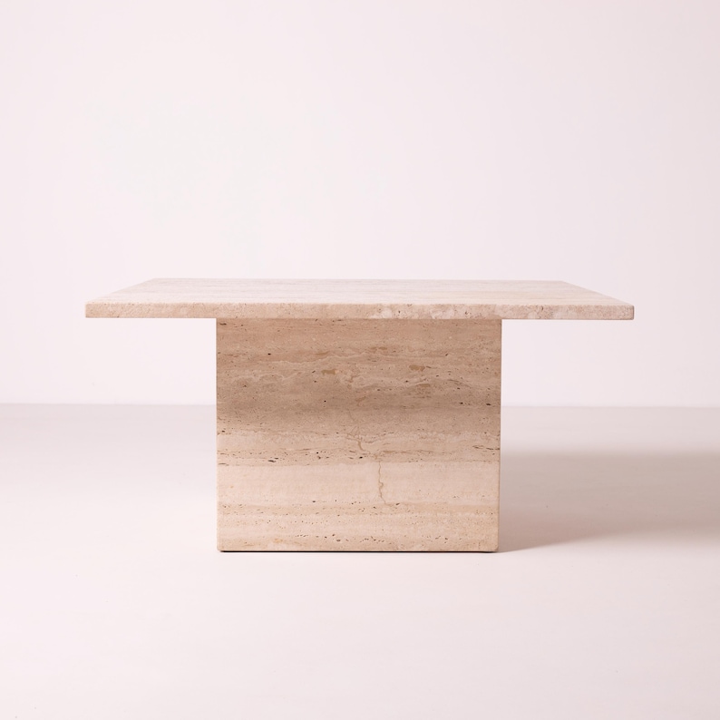 Pu&ograve; includere: Un tavolino quadrato in travertino beige. Il tavolo ha una base rettangolare solida e un piano piatto. La pietra naturale ha un aspetto stratificato e strutturato. Dimensioni approssimative: 100 cm x 100 cm.