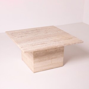 Pu&ograve; includere: Un tavolino quadrato in travertino beige chiaro. Il tavolo ha una base solida a forma di blocco e un piano quadrato e piatto. La pietra naturale presenta sottili venature e variazioni di colore. Misura circa 90 cm x 90 cm. Adatto per un soggiorno.