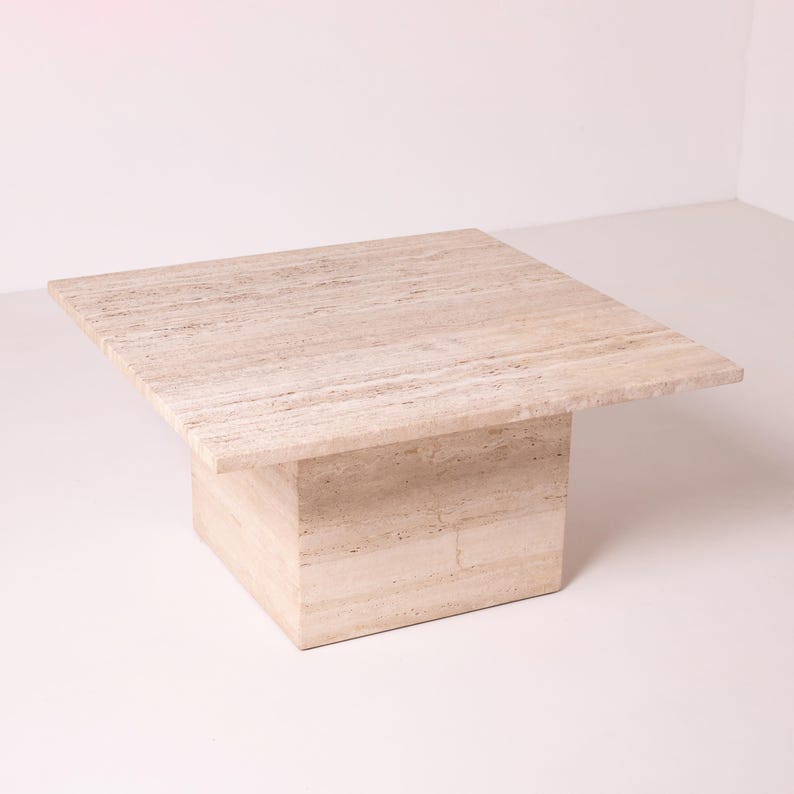 Pu&ograve; includere: Un tavolino quadrato in travertino beige chiaro. Il tavolo ha una base solida a forma di blocco e un piano quadrato e piatto. La pietra naturale presenta sottili venature e variazioni di colore. Misura circa 90 cm x 90 cm. Adatto per un soggiorno.
