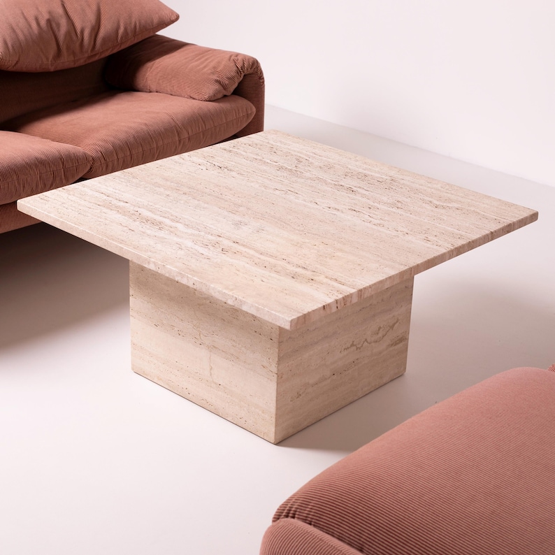 Pu&ograve; includere: Un tavolino quadrato in travertino beige con una base solida a forma di blocco. Il piano del tavolo presenta un motivo venato naturale. Il tavolo &egrave; posizionato di fronte a un divano in velluto a coste e una poltrona abbinata, creando un salotto.
