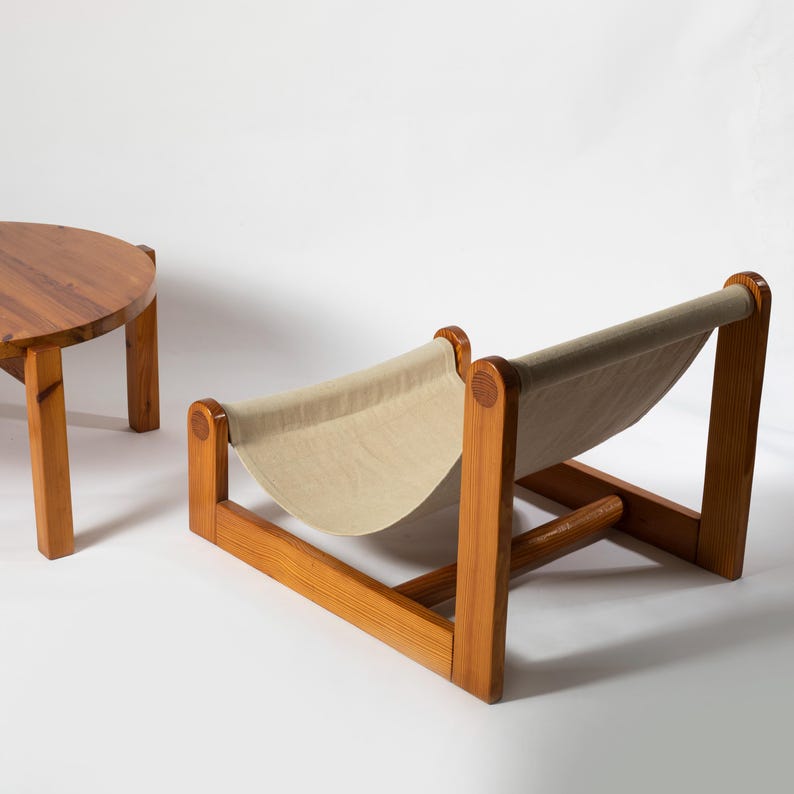 Może przedstawiać: Krzesło w stylu mid-century modern z jasnobeżowym pł&oacute;ciennym siedziskiem i drewnianą ramą. Krzesło połączone jest z okrągłym drewnianym stolikiem bocznym. Meble wykonane są z jasnego drewna i ustawione na białym tle.
