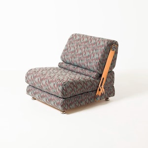 May include: A vintage armchair with a low profile, upholstered in a patterned fabric of grey, red, and teal. The chair has a layered design, with a backrest and seat. It features a light brown leather armrest and rests on small, silver feet.