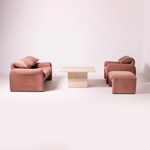 Pu&ograve; includere: Un moderno set da soggiorno in un ambiente studio. Il set comprende un divano rosa antico, due poltrone e un pouf. Un tavolino quadrato in travertino &egrave; al centro. L'arredamento ha un design morbido e arrotondato.