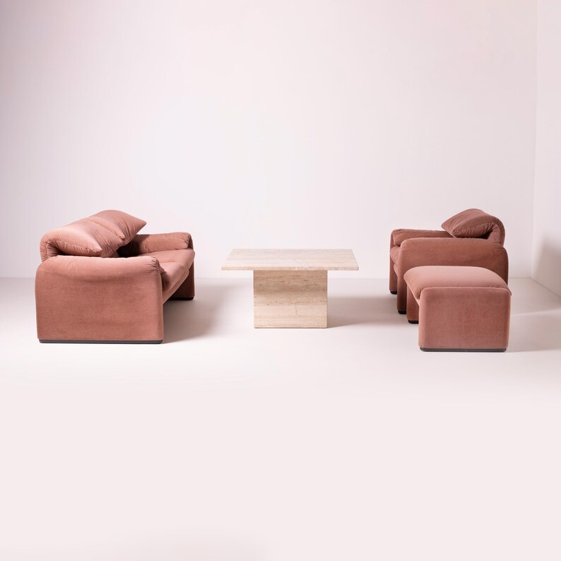 Pu&ograve; includere: Un moderno set da soggiorno in un ambiente studio. Il set comprende un divano rosa antico, due poltrone e un pouf. Un tavolino quadrato in travertino &egrave; al centro. L'arredamento ha un design morbido e arrotondato.