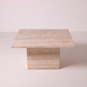 Pu&ograve; includere: Un tavolino quadrato in travertino beige. Il tavolo ha una base rettangolare solida che supporta un piano quadrato e piatto. La pietra naturale ha un motivo a strisce e una palette di colori neutri.