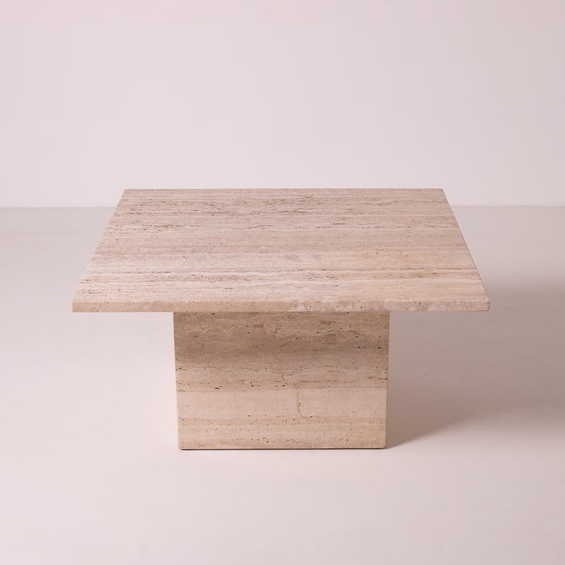 Pu&ograve; includere: Un tavolino quadrato in travertino beige. Il tavolo ha una base rettangolare solida che supporta un piano quadrato e piatto. La pietra naturale ha un motivo a strisce e una palette di colori neutri.