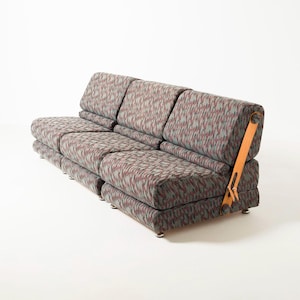 May include: A modular sofa upholstered in a patterned fabric of grey, red, and green hues. The sofa has a low design with individual seat cushions and backrests. It features a tan leather strap detail and rests on small, silver feet.