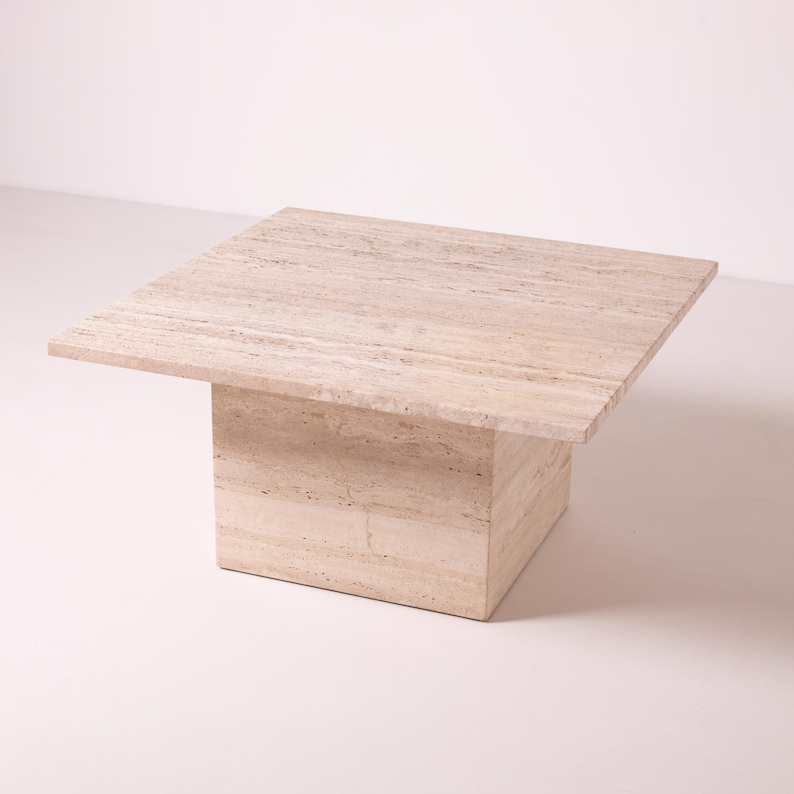 Pu&ograve; includere: Un tavolino quadrato in travertino beige. Il tavolo ha una base rettangolare solida che supporta un piano quadrato e piatto. La pietra presenta venature naturali e una palette di colori neutri, adatta a vari stili di interni.