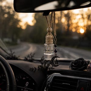 May include: A clear glass car air freshener with a wooden top and a decorative charm featuring a crescent moon and star hangs from the rearview mirror. The charm is attached to a beaded chain. The car interior is visible.