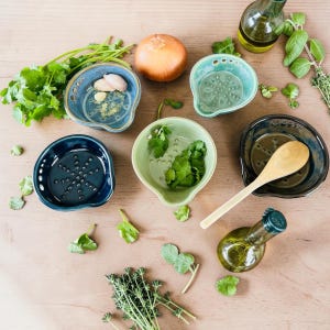 May include: A collection of five ceramic grater bowls in shades of blue and green, featuring textured centers for grating. The set includes a wooden spoon and is surrounded by fresh cilantro, a whole onion, and two glass bottles of olive oil.
