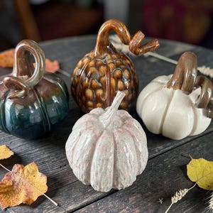 May include: Four ceramic pumpkin figurines with glossy finishes. One is dark blue and green, one is brown with a textured pattern, one is cream-colored, and one is a speckled light pink. Each features a curved, glazed stem on top.