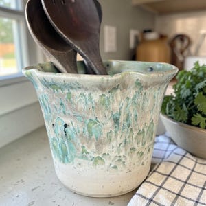 Könnte beinhalten: Ein Keramik-Utensilienhalter mit weißem Sockel und türkisfarbener und grauer, gesprenkelter Glasur. Zwei hölzerne Salatbestecke befinden sich im Halter. Der Halter steht auf einer Arbeitsplatte neben einer Schüssel mit Kräutern und einem karierten Tuch.