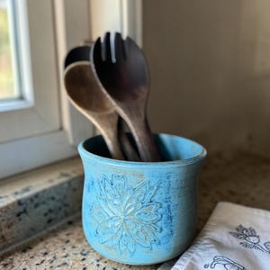 May include: A light blue ceramic utensil holder with a floral design, holding wooden spoons. The holder has a distressed finish and sits on a speckled countertop. A white dish towel with a floral pattern is in the background.