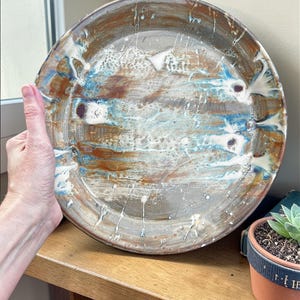 May include: A large, handmade ceramic plate with a unique, abstract design. The plate features a blend of brown, blue, and white colors, with a textured surface. The plate is held by a hand, showcasing its size and craftsmanship. A decorative piece for the home.