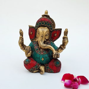 May include: A gold-colored Ganesha statue with red and green accents. The statue is sitting on a white surface with rose petals scattered around it.